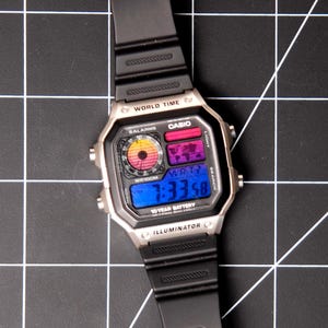 May include: A silver Casio digital watch with a black band. The watch face displays the time, world map, and other functions. The watch has the text "WORLD TIME", "CASIO", and "ILLUMINATOR".
