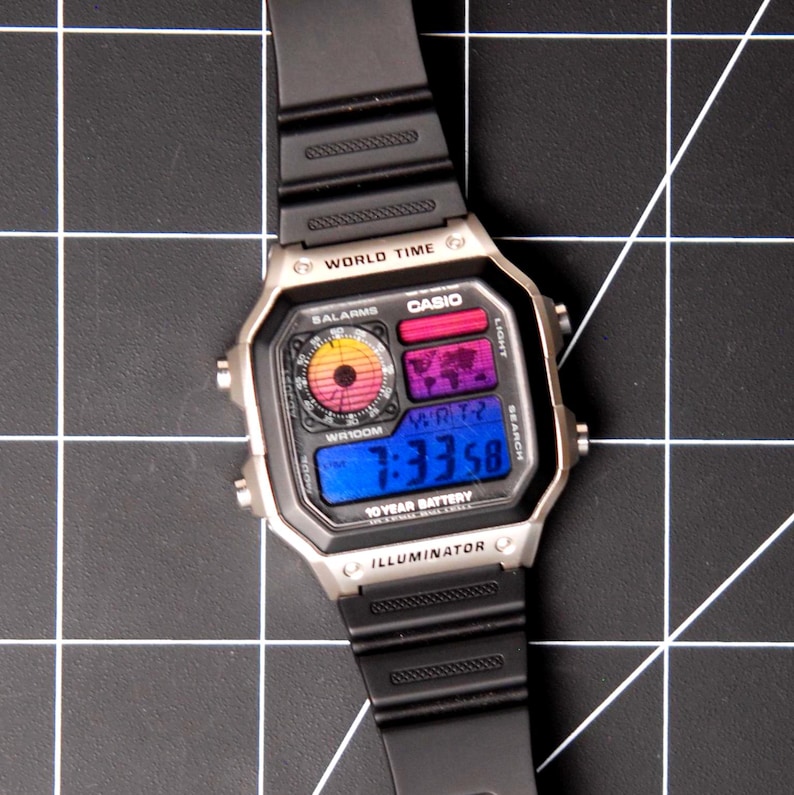 May include: A silver Casio digital watch with a black band. The watch face displays the time, world map, and other functions. The watch has the text "WORLD TIME", "CASIO", and "ILLUMINATOR".