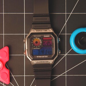 May include: A black digital watch with a silver-colored face and black band. The watch displays the time and has the text "CASIO" and "ILLUMINATOR". A red and pink fidget spinner and a blue object are also in the image.
