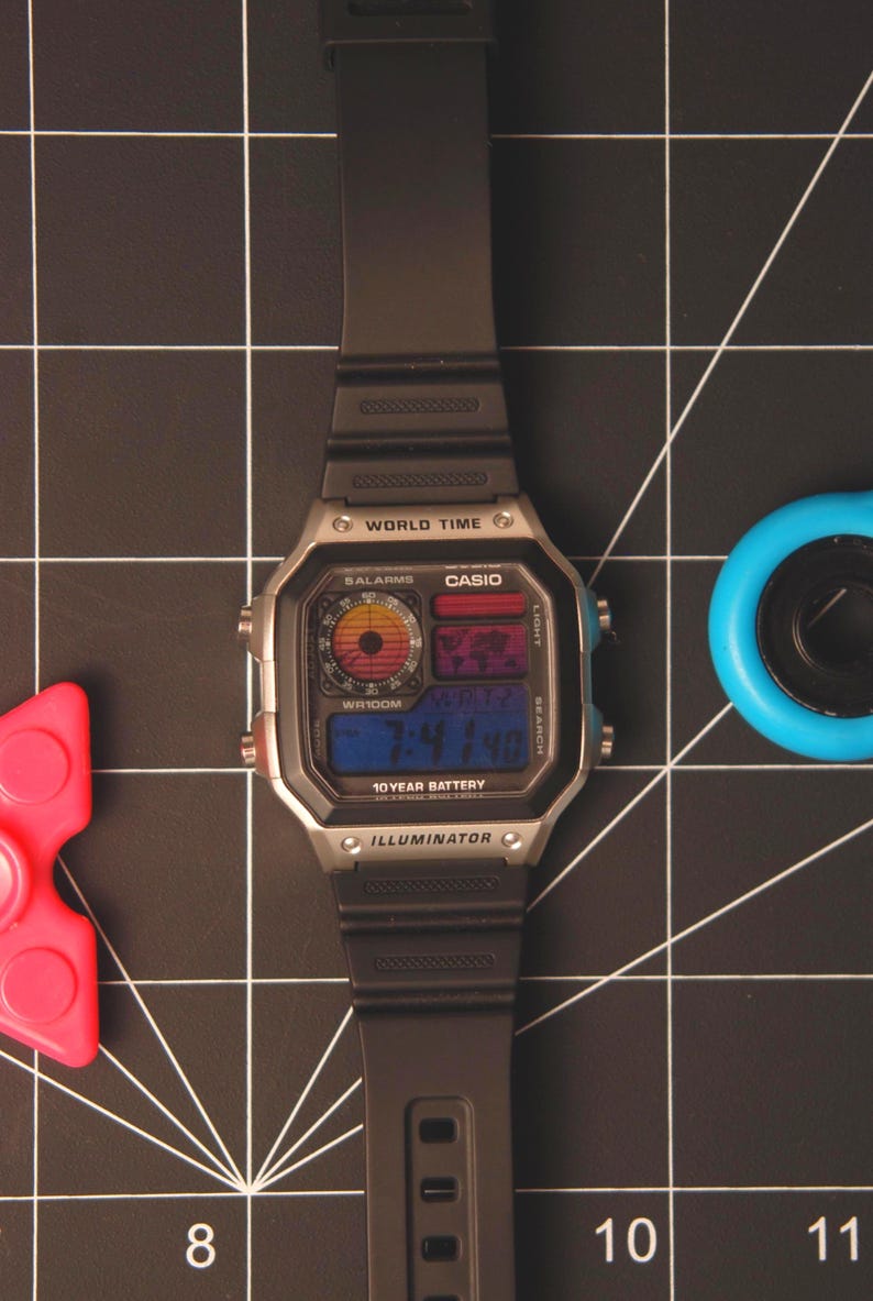 May include: A black digital watch with a silver-colored face and black band. The watch displays the time and has the text "CASIO" and "ILLUMINATOR". A red and pink fidget spinner and a blue object are also in the image.