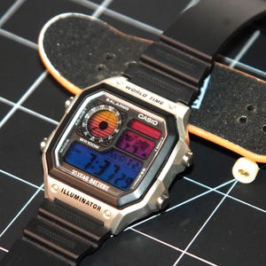 May include: A silver Casio digital watch with a black band, featuring a multi-colored display showing the time. The watch is next to a miniature skateboard on a black grid surface. The watch face displays "WORLD TIME" and "ILLUMINATOR".