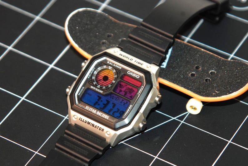 May include: A silver Casio digital watch with a black band, featuring a multi-colored display showing the time. The watch is next to a miniature skateboard on a black grid surface. The watch face displays "WORLD TIME" and "ILLUMINATOR".
