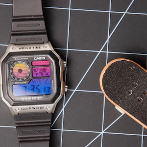 May include: A silver Casio digital watch with a black band and a small Tech Deck skateboard on a black grid surface. The watch face displays the time and world time features. The skateboard has the text "TECH DECK".
