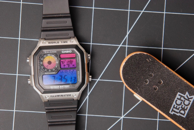 May include: A silver Casio digital watch with a black band and a small Tech Deck skateboard on a black grid surface. The watch face displays the time and world time features. The skateboard has the text "TECH DECK".