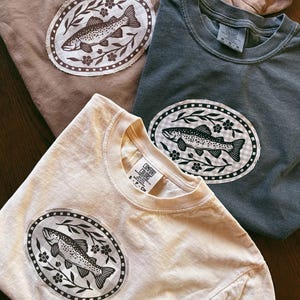 May include: Three crew neck t-shirts in tan, charcoal gray, and cream. Each shirt features a central oval patch with a dark brown or black graphic of a fish surrounded by floral vines and a checkered border. The shirts have Comfort Colors labels.