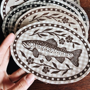 May include: A stack of oval fabric patches featuring a brown gingham background and a dark brown printed design of a trout fish surrounded by floral branches. Each patch is framed by a dark brown border with a repeating square pattern.