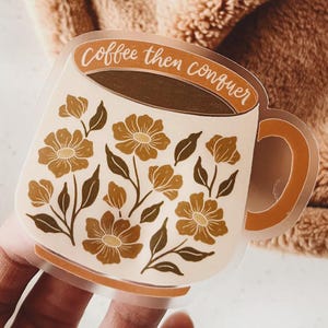 以下が含まれることがあります： 「Coffee then conquer」の文字が入ったコーヒーカップのデザインのクリアステッカー。カップにはコーヒーが入り、茶色とベージュの色合いの花柄で装飾されています。カップには茶色の取っ手が付いています。