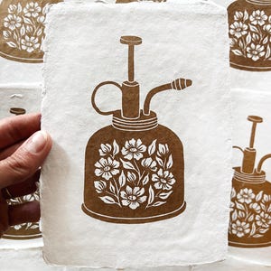 May include: Brown and white floral print of a vintage plant mister on textured paper. The mister features a bulbous body, a handle, and a spray nozzle. The floral design is a cluster of flowers and leaves.