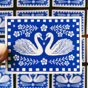 May include: A blue and white sticker featuring two swans facing each other, forming a heart shape. The design includes floral and leaf accents, with a border of white squares. The sticker is held by a hand.
