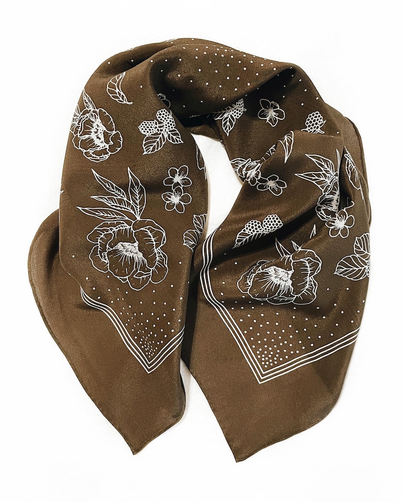 K&ouml;nnte beinhalten: Ein braunes Seidentuch mit wei&szlig;em Blumenmuster. Das Tuch hat ein sich wiederholendes Muster aus Blumen und Bl&auml;ttern mit einem gepunkteten Rand. Das Tuch ist gefaltet und zeigt seine Form und sein Design.