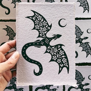 May include: A dark green dragon illustration with floral wing details and a crescent moon. The dragon is printed on textured, off-white paper. The image is held by a hand, with other similar prints visible in the background.