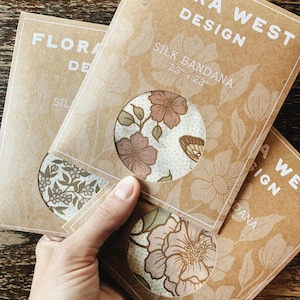 K&ouml;nnte beinhalten: Drei Seiden-Bandanas in brauner Verpackung mit floralen Designs. Die Verpackung tr&auml;gt die Aufschrift "FLORA WEST DESIGN" und "SILK BANDANA 23" x 23"". Die Bandanas weisen verschiedene Blumenmuster in Rosa-, Gr&uuml;n- und Braunt&ouml;nen (ca. 58 cm x 58 cm) auf.