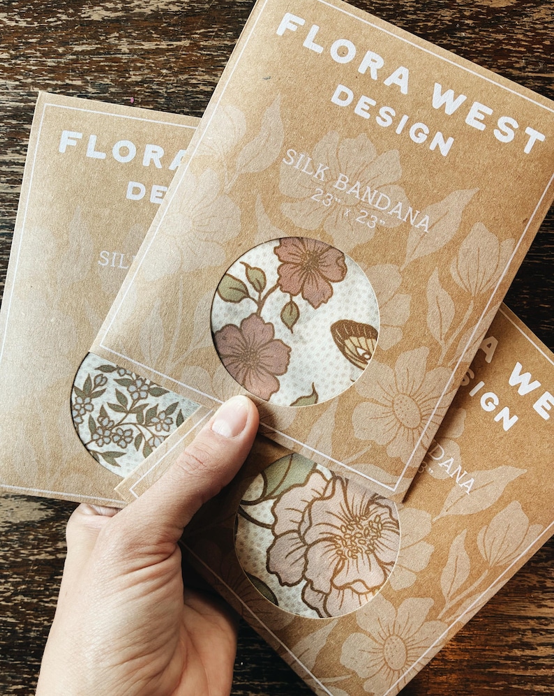 K&ouml;nnte beinhalten: Drei Seiden-Bandanas in brauner Verpackung mit floralen Designs. Die Verpackung tr&auml;gt die Aufschrift "FLORA WEST DESIGN" und "SILK BANDANA 23" x 23"". Die Bandanas weisen verschiedene Blumenmuster in Rosa-, Gr&uuml;n- und Braunt&ouml;nen (ca. 58 cm x 58 cm) auf.
