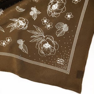 K&ouml;nnte beinhalten: Ein braunes quadratisches Tuch mit einem wei&szlig;en Blumenmuster. Das Design umfasst gro&szlig;e Blumen, Beeren und Bl&auml;tter. Das Tuch hat einen wei&szlig;en Rand und den Text "FLORA WEST DESIGN."