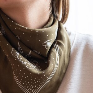 K&ouml;nnte beinhalten: Olivgr&uuml;nes Bandana mit wei&szlig;em Blumen- und Punktmuster. Das quadratische Tuch ist gefaltet und um den Hals getragen. Die R&auml;nder sind mit einem wei&szlig;en Rand umrandet. Das Bandana ist aus einem weichen, seidigen Material.