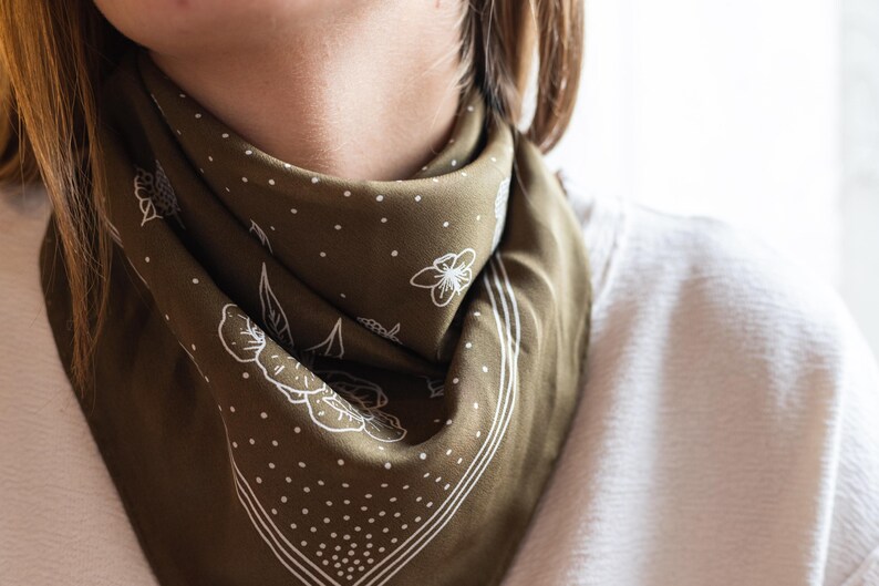 K&ouml;nnte beinhalten: Olivgr&uuml;nes Bandana mit wei&szlig;em Blumen- und Punktmuster. Das quadratische Tuch ist gefaltet und um den Hals getragen. Die R&auml;nder sind mit einem wei&szlig;en Rand umrandet. Das Bandana ist aus einem weichen, seidigen Material.