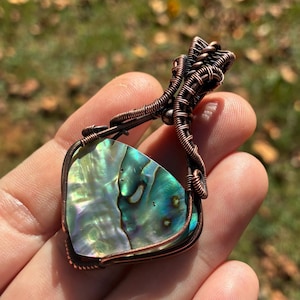 May include: A pendant featuring an iridescent abalone shell slice secured within a decorative copper wire-wrapped frame. The shell displays shifting hues of green, blue, and purple, while the dark copper wire forms a complex, coiled bail.