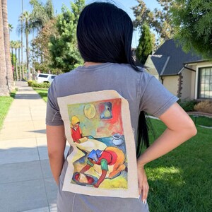 May include: A gray t-shirt with a colorful painting on the back. The painting depicts two figures in a kitchen setting, with vibrant reds, yellows, and blues. The artwork is framed by a beige fabric.