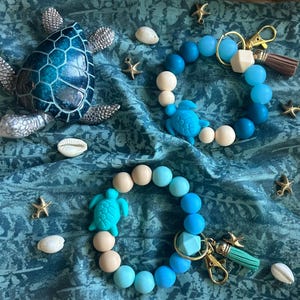 May include: Two beaded wristlet keychains with sea turtle charms in shades of blue and cream are shown. A decorative sea turtle figurine and starfish charms are scattered around the keychains. The background is a blue patterned fabric.