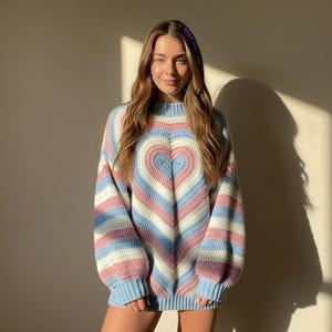 Può includere: Un maglione dai colori pastello con un grande cuore sul davanti. Il maglione presenta strisce orizzontali in azzurro, rosa e bianco. Il cuore è centrato e delineato in bianco. Le maniche e l'orlo sono di un azzurro chiaro uniforme.