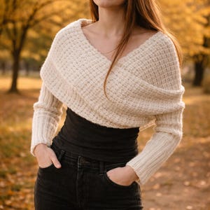 Puede incluir: Bolero de punto color crema con mangas largas. El diseño envolvente presenta un escote en V profundo y se usa sobre una camiseta negra y vaqueros oscuros. El fondo muestra una escena otoñal borrosa con follaje amarillo y marrón.