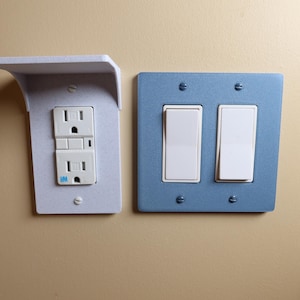 Peut inclure: Deux plaques murales électriques. L'une est blanche avec une double prise, l'autre est bleue avec deux interrupteurs. Les deux sont montées sur un mur beige. La plaque de prise a une petite étagère.