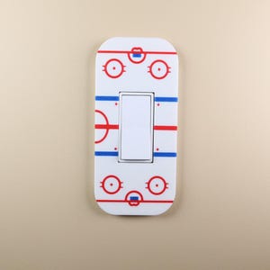 May include: White light switch cover with a hockey rink design. It features red and blue lines and circles, and a rectangular switch in the middle. A decorative item for a hockey enthusiast's home.