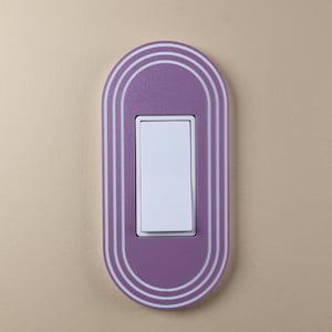 May include: A lavender light switch cover with a white rectangular switch. The cover has a rounded rectangular shape with three white concentric lines. The switch is centered within the cover. The design adds a modern touch to any room.