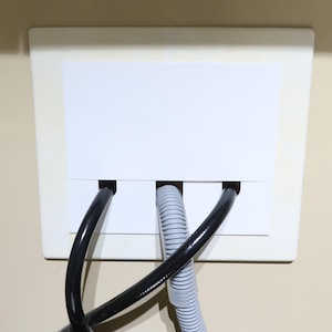Peut inclure: Une plaque murale rectangulaire blanche avec trois ouvertures, chacune abritant un câble. Deux câbles noirs encadrent un câble gris ondulé. La plaque est fixée sur un mur beige.
