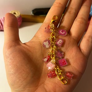 May include: A gold-toned metal charm bracelet featuring multiple small, translucent acrylic charms in shades of pink and red. The charms include various shapes such as a cat silhouette, a letter C, the number 2, and several stylized character figures.
