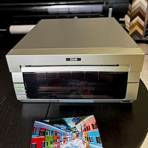 May include: Silver DS40 photo printer with a black front panel and control buttons. A colorful print of buildings with blue skies and clouds is on the table. The printer is on a dark surface.
