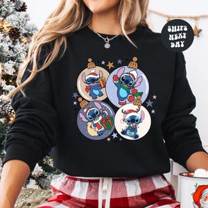 A woman wearing a black sweatshirt with a Christmas-themed design featuring four cartoon characters, including Stitch, surrounded by ornaments and stars.