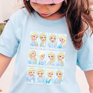 A young girl wearing a light blue t-shirt with a collage of cartoon images of a blonde girl with various facial expressions and emotions.