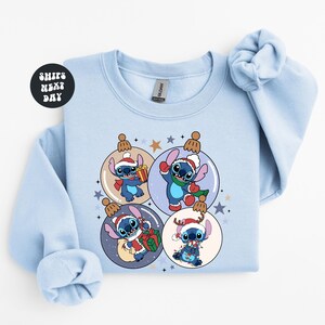 A light blue sweatshirt with a Christmas-themed design featuring four cartoon characters from the Lilo & Stitch movie franchise, each wearing a unique outfit and surrounded by festive ornaments.