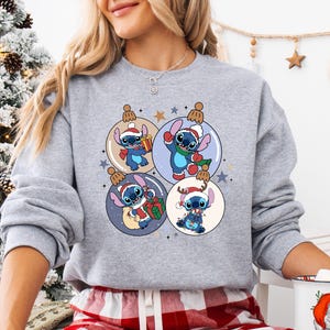 A woman wearing a gray sweatshirt with a Christmas-themed graphic design featuring four cartoon characters, including Stitch, surrounded by ornaments and stars.
