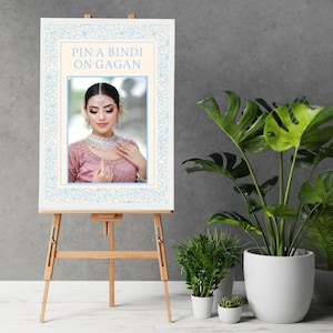 May include: A wooden easel displays a sign with the text "PIN A BINDI ON GAGAN." The sign features a photo of a person wearing a pink outfit and jewelry. The sign has a light blue floral border. Potted plants are in the background.