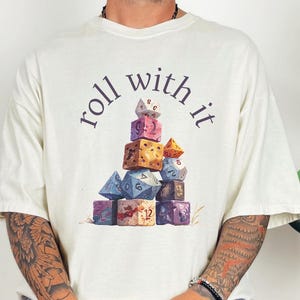 Puede incluir: Camiseta color crema con la frase "roll with it" sobre una pila de dados coloridos. Los dados son de varias formas y colores, incluyendo azul, morado, marrón y rosa. La camiseta la lleva una persona.