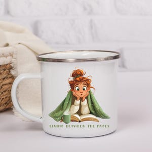 May include: White enamel mug with a silver rim and handle. The mug features an illustration of a girl wrapped in a green blanket, reading a book. Text on the mug reads "LIVING BETWEEN THE PAGES". A small potted plant and a woven basket are in the background.