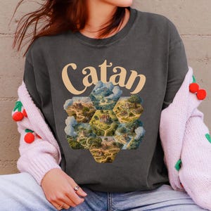 May include: Dark gray t-shirt with the word "Catan" in a light yellow font. The shirt features a colorful hexagonal graphic. The person is wearing light blue jeans, green sneakers, and a pink cardigan with red cherry accents.