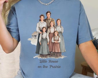 May include: A light blue t-shirt featuring a graphic of a family from the Little House on the Prairie series. The graphic includes the text "Little House on the Prairie" in a script font. The shirt is a crew neck style.
