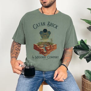 May include: Olive green t-shirt with the text "CATAN Brick & Masonry Company" and a graphic of a brick structure with a river and geometric shapes. The shirt is worn with blue jeans.