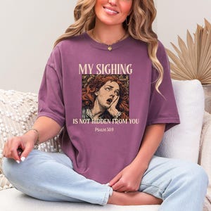 Può includere: T-shirt color malva con la scritta "MY SIGHING IS NOT HIDDEN FROM YOU" e "PSALM 38:9". La maglietta presenta un'illustrazione vintage di una donna dai capelli rossi. La maglietta ha una vestibilità ampia.