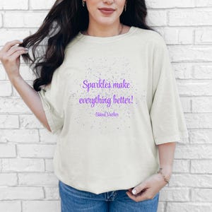 Op de afbeelding: Crèmekleurig T-shirt met de paarse tekst "Sparkles make everything better!". Het shirt heeft een losse pasvorm en wordt gecombineerd met een blauwe jeans. De quote is van Bikni Vacker.