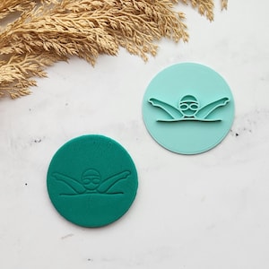 May include: Two circular cookie stamps, one teal and one dark teal, each depicting a swimmer in a freestyle stroke, wearing goggles. The stamps are on a white surface with dried plants in the background.