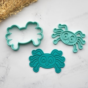 May include: Three teal-colored spider-shaped cookie cutters. One is the outline of a spider, and the other two are detailed spider shapes with smiling faces. The cutters are made of plastic and are on a white marble surface.