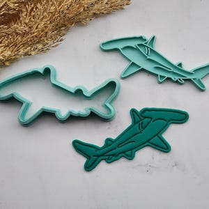 Peut inclure: Trois emporte-pièces en forme de requin marteau de couleur sarcelle et un contour sarcelle d'un emporte-pièce en forme de requin marteau sont présentés sur une surface blanche. Les emporte-pièces sont en plastique et de tailles variées. Des épis de blé séchés sont en arrière-plan.