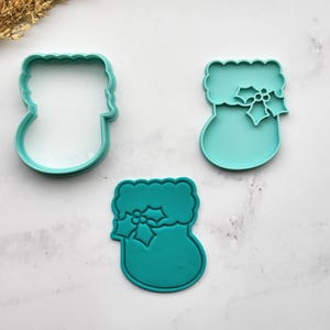 May include: Three teal plastic cookie cutters shaped like Christmas stockings. Two have a holly leaf and berry design. The cutters are on a white marble surface.