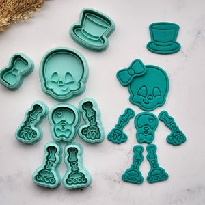 May include: A collection of teal-colored cookie cutters in the shape of a skeleton, including a skull, bow tie, top hats, and bone-shaped limbs. The cutters are arranged on a light-colored surface, ideal for Halloween or themed baking.
