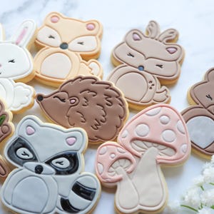 Könnte beinhalten: Eine Sammlung dekorierter Zuckerkekse in Form von Waldtieren. Die Kekse umfassen ein weißes Kaninchen, einen Fuchs, ein Reh, einen Bären, einen Igel, einen Waschbären, einen Baumstumpf und Pilze. Die Kekse sind mit verschiedenen Farben und Details verziert.