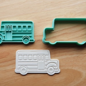May include: Three school bus-shaped cookie cutters in different styles. One is a solid teal color, another is a teal outline, and the third is a white embossed design. The cutters are made of plastic and sit on a wooden surface.
